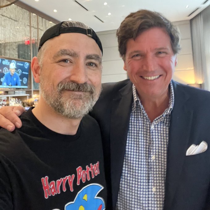 Two men smile for a selfie in what appears to be a restaurant or lounge setting, with one wearing a black cap and a "Harry Potter" t-shirt (also featuring blue cartoon character imagery) and the other dressed in a navy blazer with a checkered shirt and white pocket square, identified by the post as Tucker Carlson.