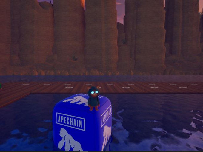 A virtual penguin character with glowing blue eyes stands on a blue 'APECHAIN' branded cube floating in water, set against a backdrop of tall reddish cliffs in a VR environment.