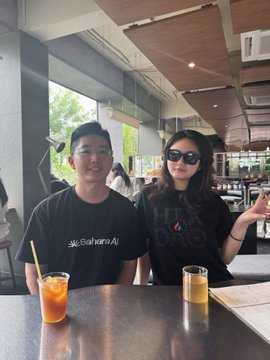 Two young people sit at a table in a cafe; the man wears a black 'Sahara AI' t-shirt while the woman wears sunglasses and a black shirt, with orange and yellow drinks in front of them.