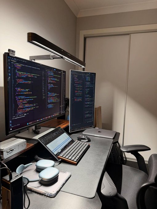 A dual-monitor coding setup featuring a curved ultrawide monitor and a vertical monitor displaying code, accompanied by a mechanical keyboard, laptop, headphones, and an ergonomic chair.
