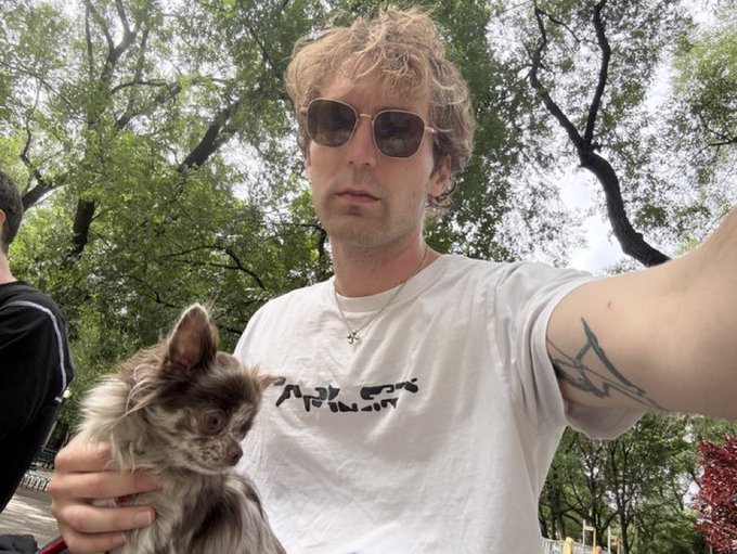 A man wearing sunglasses and a white t-shirt holds a small Chihuahua dog in an outdoor setting with trees in the background.