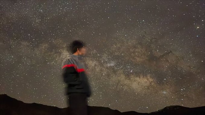 An astrophotography shot capturing the Milky Way galaxy stretching across the night sky, with a person in a striped jacket silhouetted in the foreground looking up at the stars.