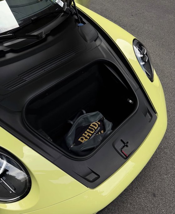 A yellow sports car with its hood open to reveal a front trunk compartment, which contains a black bag with 'RHUDI' printed in gold lettering.
