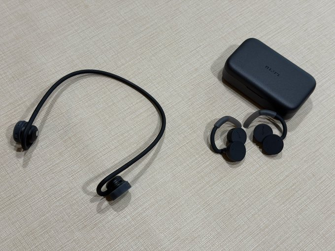 Black bone-conduction style wireless headphones with a neckband alongside their charging case and earbud accessories on a beige textured surface.