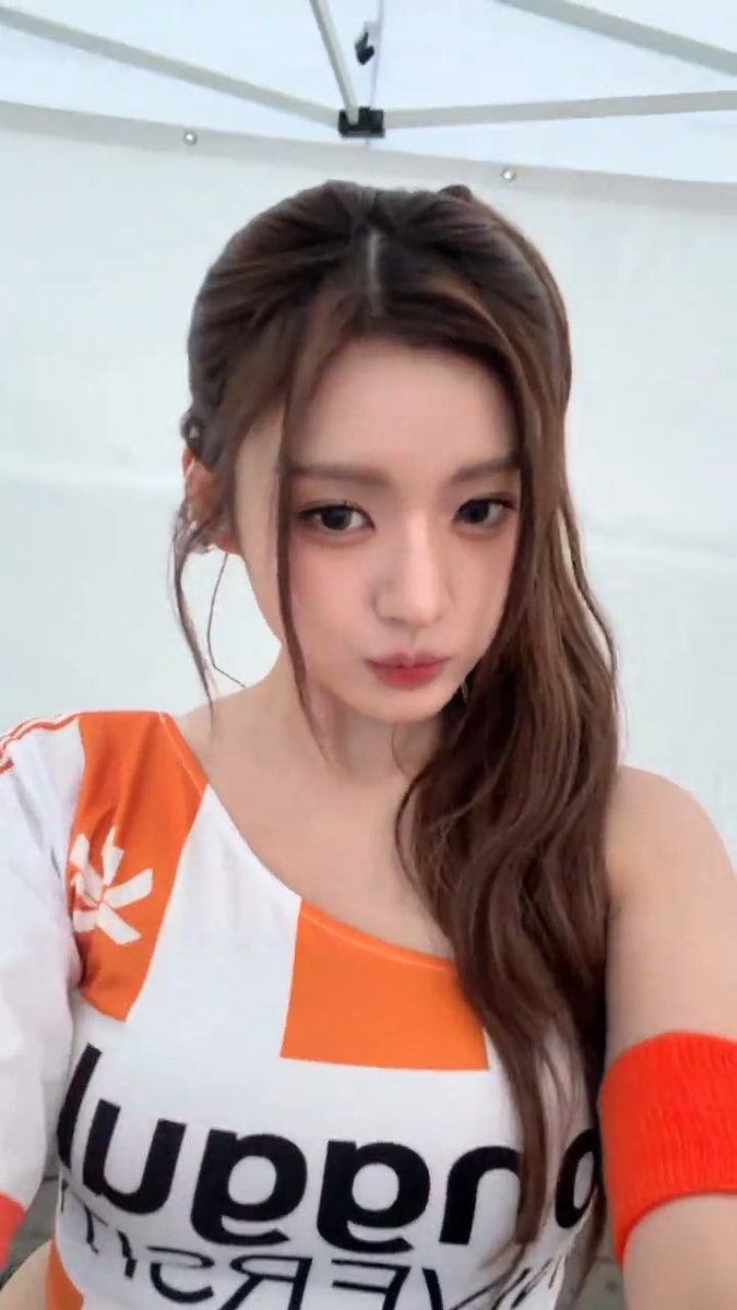 Close-up selfie of a young Asian woman with brown hair wearing an orange and white off-shoulder top, with "SUPER" text visible and an orange arm sleeve.
