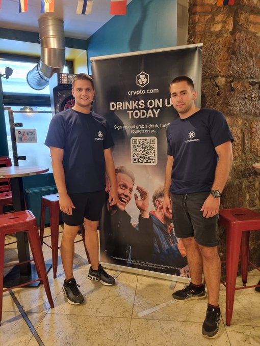 The image shows two men standing next to each other with their arms around each other's shoulders. They are both wearing blue shirts and black shorts. Behind them, there is a banner that says "Drinks on us today" and has a QR code on it. The banner also mentions the website "crofam.com". One of the men is holding a drink in his hand while they pose for the photo.
