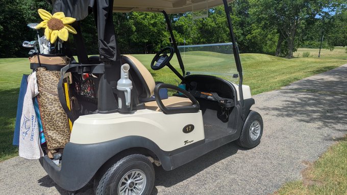 The image shows a golf cart parked next to a road, with several golf clubs hanging from its roof. The golf cart has a white exterior and black interior, giving it an elegant appearance. There are two people visible in the scene - one standing near the left side of the image and another person closer to the right side. A yellow flower is also present on the golf cart's roof.

The post that accompanies this image reads: "#Q #Golf #xrp #0doubt". This post seems to be related to a discussion or review about golf, possibly featuring the golf cart and its surroundings.
