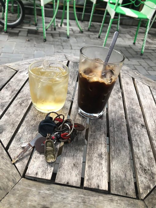 Two iced beverages sit on a round wooden table next to a set of keys, with green metal chairs visible in the background.
