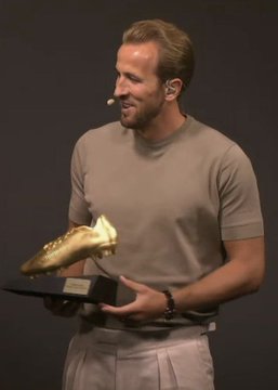 A man in a beige t-shirt and light pants standing on a presentation stage, holding a golden boot-shaped trophy.
