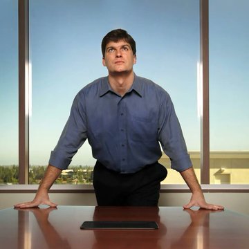 A stock photo of a Middle Eastern man in a blue button-up shirt sitting at a desk, leaning forward with a shocked, wide-eyed expression.