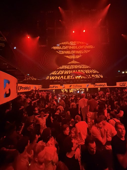 A packed crypto party bathed in red lighting, featuring a large screen displaying repeating "BLOFIN" and "WHALES" logos, alongside brands such as "Probly," "TxFlow," and "Bitget." The energetic crowd captures the fire vibe of the event.