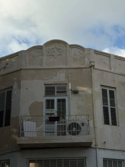 A photo of an old, weathered building facade featuring a Star of David decorative emblem at the top, with shuttered windows and a small balcony.