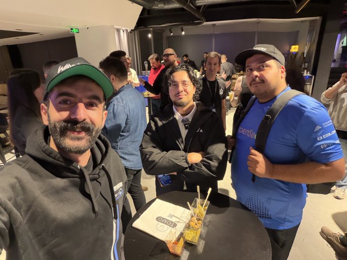 Three men pose at a networking event, with the table in front displaying an "OWO" branded document, one of whom wears a "PenPen" branded shirt featuring cryptocurrency-related logos, suggesting a Web3 gaming industry gathering where the poster likely connected with fellow attendees to support emerging projects.