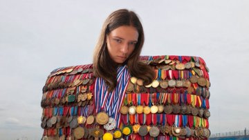 A person with long brown hair wears an oversized jacket covered in colorful ribbons and medals, with a medal hanging around their neck, standing against a blurred background.