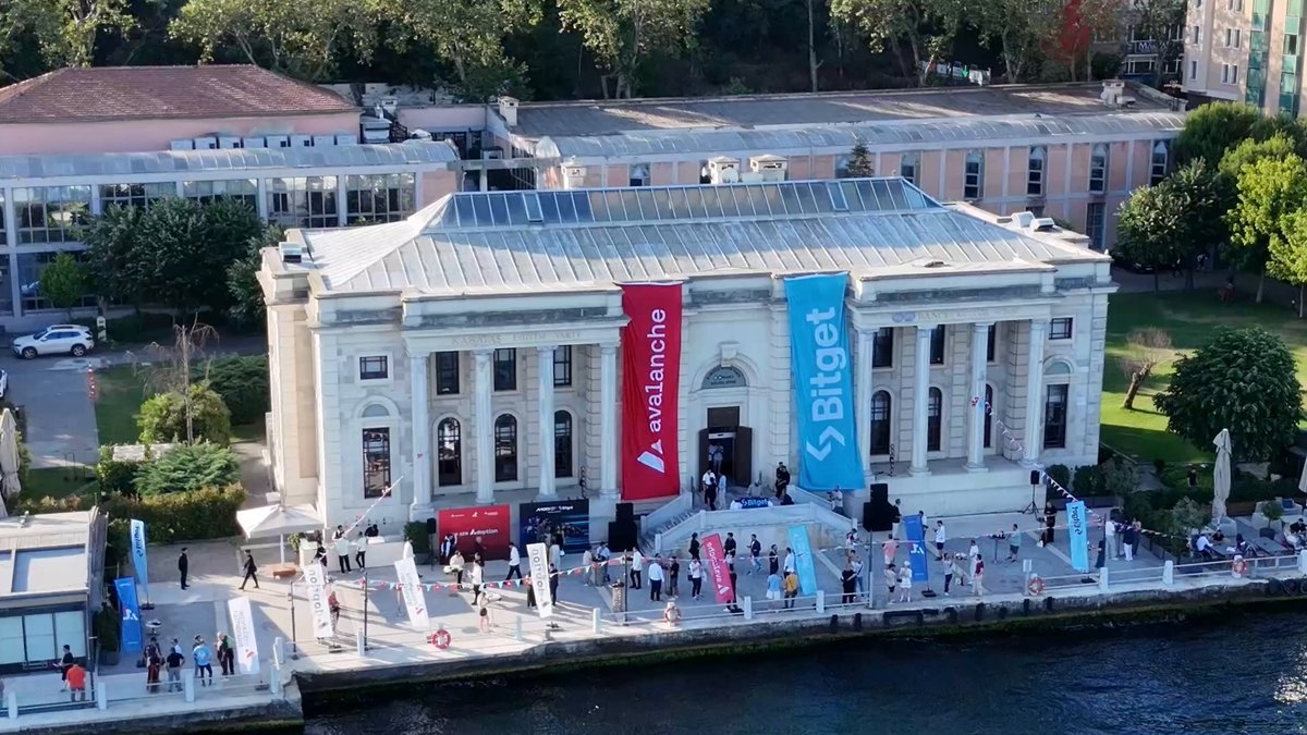 The image shows an aerial view of a large building, possibly a museum or a historical site, situated next to a body of water. The building has banners hanging from its roof, indicating that there might be an event taking place inside. There are several people gathered around the building and near the water's edge, suggesting they have come together for some sort of gathering or celebration. The post also mentions "Team1 Crypto House" and "@bitget", which could possibly be related to a specific event or exhibition happening at the location.