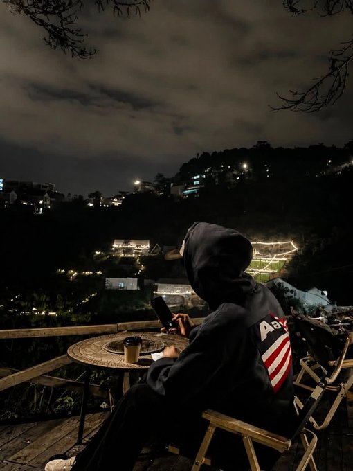 A person wearing a dark hoodie with an American flag graphic sits at a wooden table with a coffee cup, looking at their phone at night overlooking a city.