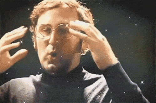 A close-up shot of a man with short brown hair and wire-rimmed glasses, wearing a dark shirt. He has his hands raised on either side of his head and his mouth is open as if speaking or making an 'o' shape, with small sparkling star effects overlaid on the footage.