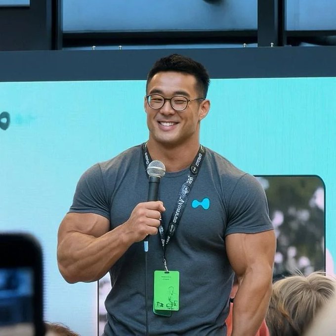 A photo of a smiling, muscular man wearing glasses and a gray t-shirt with a small logo, holding a microphone and wearing a lanyard with a green ID badge that appears to say 'CREATOR', speaking at what looks like a conference or panel.