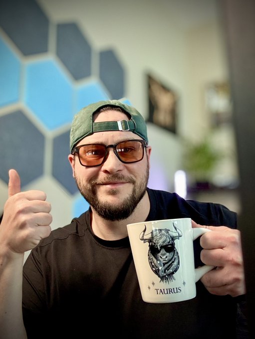 The image features a man wearing sunglasses, a black shirt, and a green baseball cap holding a white mug with a design of a Taurus symbol on it. The caption reads "GM Tuesday! Quick programming note here, stream at 2pm ET for today. Let'S get after it fam; keep crushing it and looking forward to the @SolOnChain live stream in a couple hours with some REAL LEGENDS!".