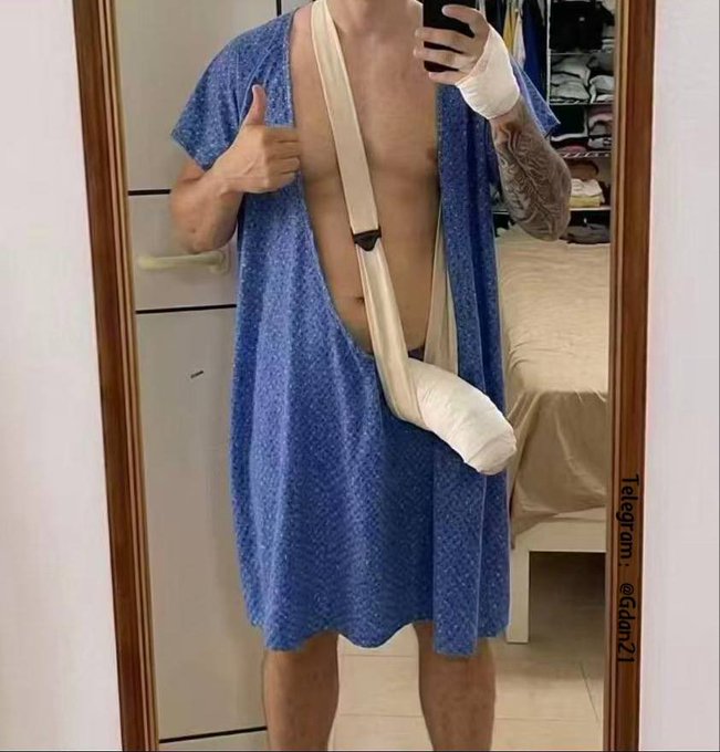 A mirror selfie shows a person in a blue hospital gown with a bandaged hand and arm sling, making a thumbs-up gesture.