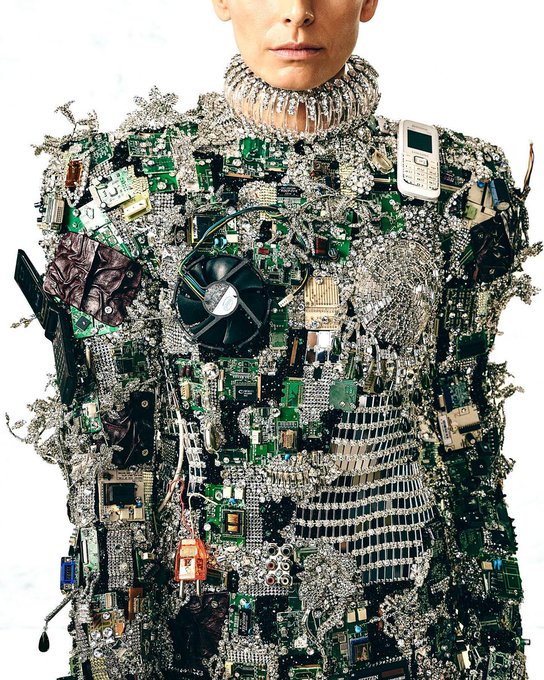 A model wears an avant-garde couture garment covered entirely in electronic waste, featuring circuit boards, a computer cooling fan, a Nokia mobile phone, tangled wires, and crystals against a white background.