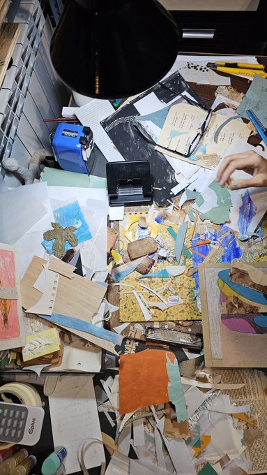 A cluttered art workspace is strewn with scraps of colorful waste paper, cardboard pieces, and painted fragments, illuminated by a black desk lamp, reflecting the artist's lifelong fascination with transforming discarded paper into creative characters. A hand reaches into the chaotic scene to sort through the materials, alongside tools like a blue paper punch and scissors that aid in the craft.