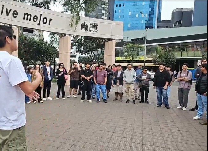 A person in a white t-shirt and green pants addresses a crowd of onlookers gathered outside a building with signs reading "ive mejor" and "SCAPE."