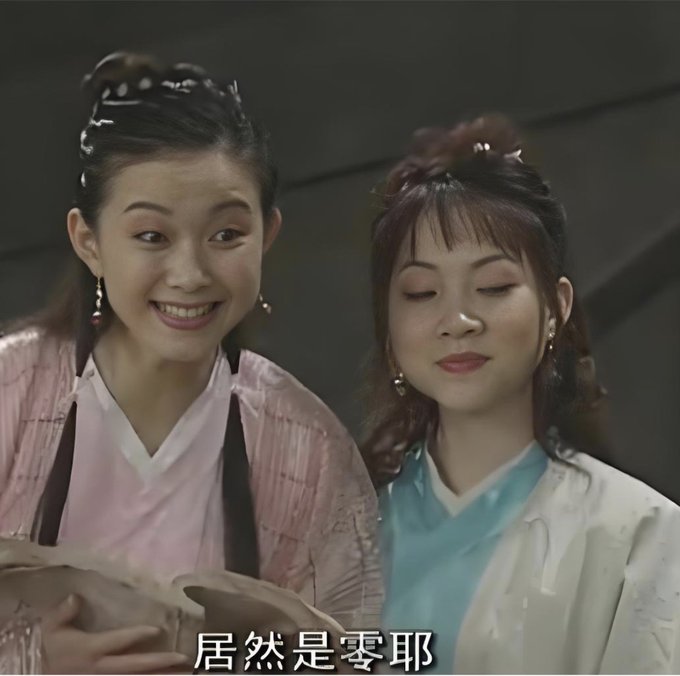 The image features two women dressed in traditional Chinese clothing standing next to each other, possibly discussing something or posing for a photo. The background of the image is dark, which contrasts with the bright colors of their outfits. There are no texts present in the image, and the focus remains on the two women and their interaction.