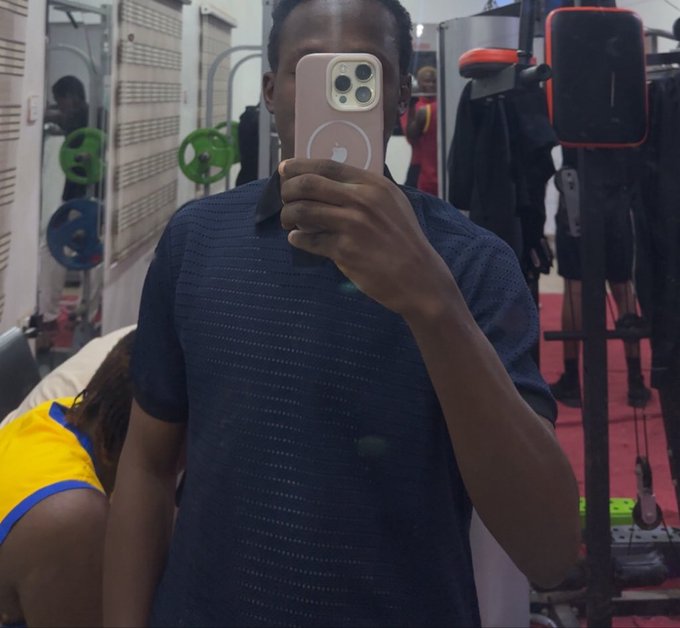 A person in a dark blue shirt takes a mirror selfie with a pink smartphone (showing the Apple logo) in a gym setting with weight racks and equipment visible.