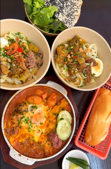 A spread of Vietnamese dishes including two bowls of bún topped with meat, peanuts, and herbs, a dish of bún chả in tomato sauce with a fried egg, a side of fresh greens, and a baguette.