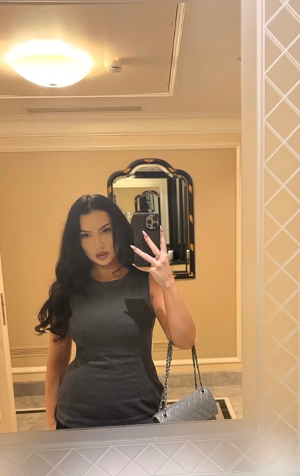 A mirror selfie of a woman with long dark hair wearing a form-fitting gray dress, taking the photo with an iPhone and holding a light blue quilted handbag in an elegantly decorated room.
