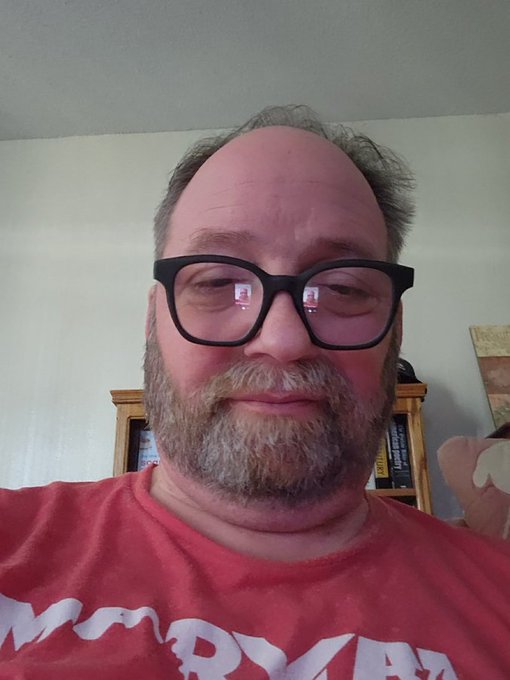 A close-up selfie of a balding man with glasses and a reddish-brown beard wearing a red t-shirt with partial text visible, taken in front of a bookshelf.