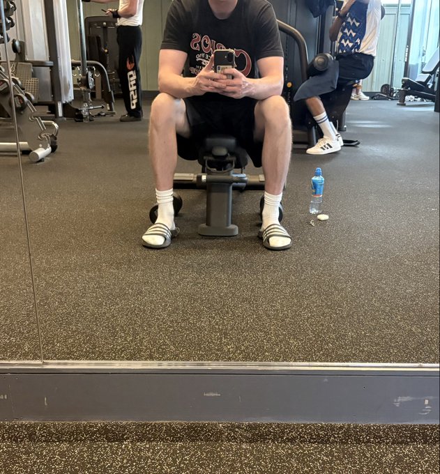 The image shows a man sitting on an exercise machine, wearing white socks and holding his cell phone. There are other people present in the gym as well, indicating that this is a public fitness facility. The caption of the image reads "We are so back Getting ready for hot Emin summer." This suggests that the photo was taken during the summer season when people tend to use exercise machines more often due to warmer weather and outdoor activities.

The man sitting on the machine appears to be using his phone, possibly checking messages or looking up information related to fitness or summer plans. The presence of other gym-goers in the background further emphasizes that this is a public space where people come together for various purposes, including exercise and socializing.