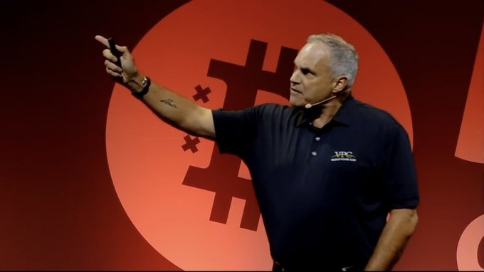 A man wearing a dark polo shirt with a "MPC" logo and a headset microphone, pointing while giving a presentation on stage with a red background featuring a large cross/sword graphic.