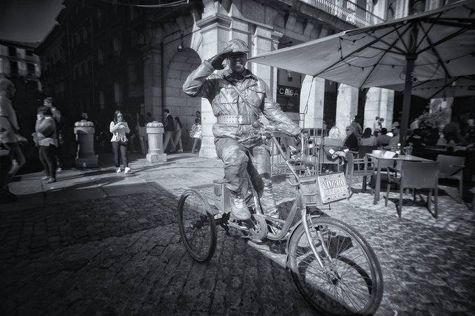 A black and white photo shows a street performer dressed as a silver robot riding a bicycle through a cobblestone plaza, with 'ALOHA' visible on the bike.