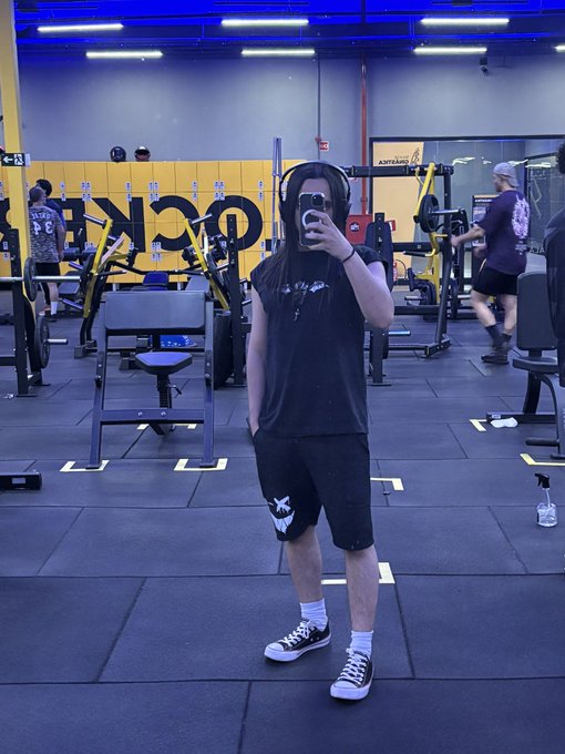 A person with long hair wearing headphones and a black sleeveless shirt takes a mirror selfie inside a gym, standing in front of rowing machines and other workout equipment.