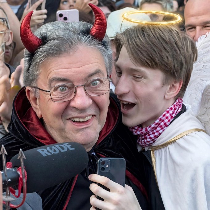 An older man wearing red devil horns and glasses being interviewed next to a younger man dressed as an angel with a halo and white wings, with a RØDE microphone visible in the foreground.