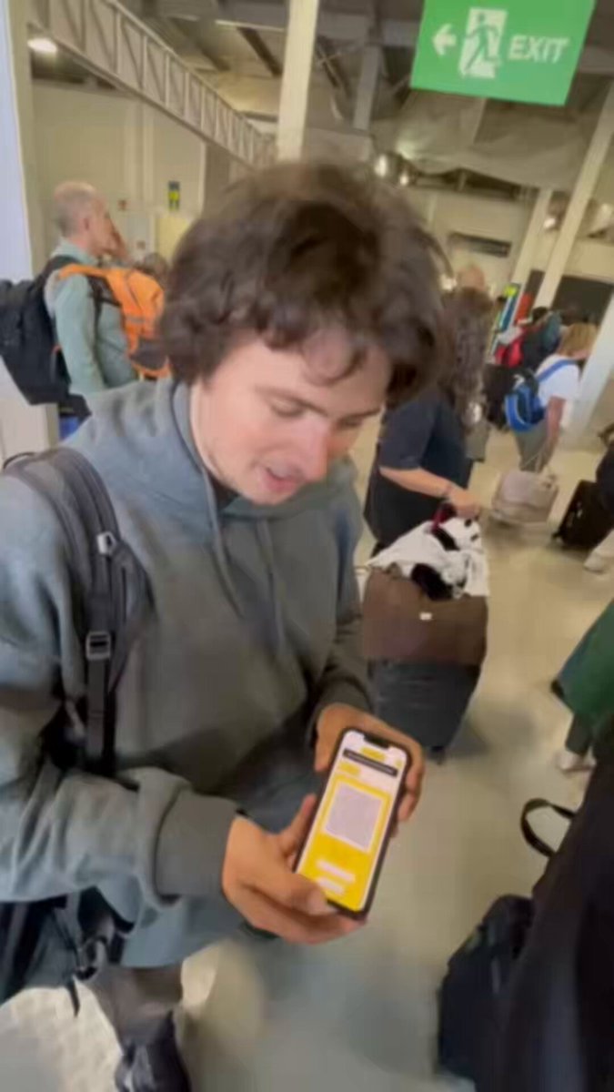 The image shows a man holding up his smartphone, which has an app that allows him to send money from his wallet to another account using the Ark lightning gateway. The man appears to be at an airport, as there are several other people around him who might also be travelers or airport staff. The post seems to be promoting a mobile payment system in Kenya for Bitcoin Cash, and it is being used by the man to transfer money from his wallet to another account.