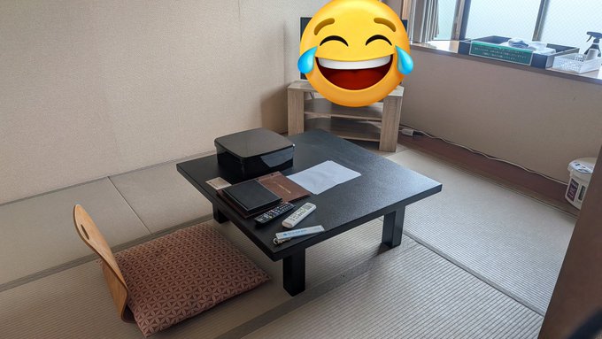 The image shows a small room with a black table, chair, and remote control placed on top of the floor. A smiley face sticker or emoticon is attached to the chair's backrest, adding a playful touch to the scene. The room appears to be empty apart from these items, creating an atmosphere that could be described as minimalistic or simple.