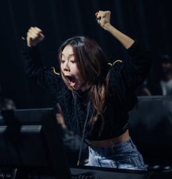 A woman with long dark hair, wearing a black textured jacket and denim shorts, enthusiastically cheers with both fists raised and mouth open in excitement.