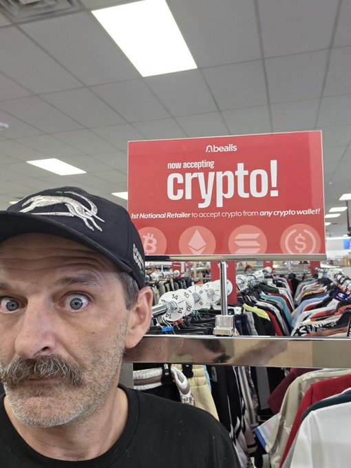 A selfie of a mustachioed man in a black cap inside a store, with a red sign behind him reading "bealls now accepting crypto! 1st National Retailer to accept crypto from any crypto wallet!" alongside Bitcoin, Ethereum, and other crypto logos.