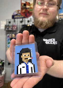 A bearded man wearing glasses and a 'GRINDER GEEKS' polo shirt holding up a pixel-art trading card or sticker depicting a character with dark hair in a white suit and blue tie.