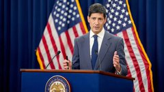 A man in a suit speaks at a podium with a presidential seal, with American flags displayed behind him.