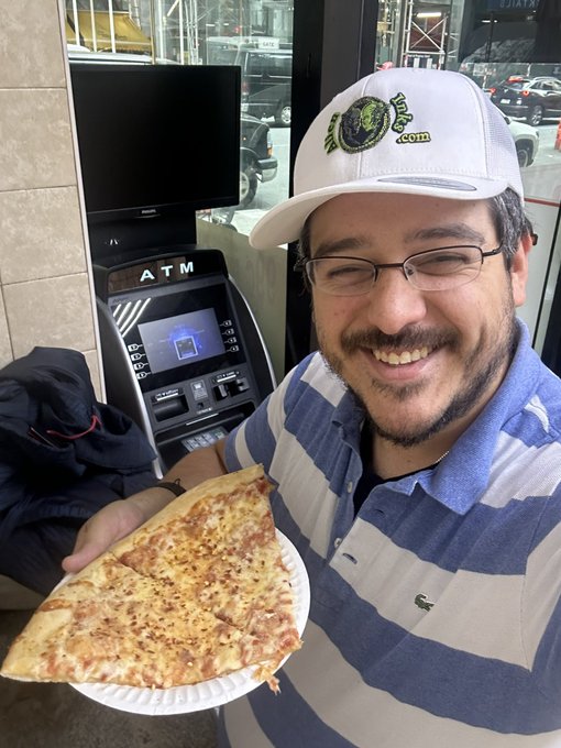 A smiling man wearing a John Deere cap, glasses, and a blue-striped Lacoste shirt holds two large slices of cheese pizza in front of an ATM machine.