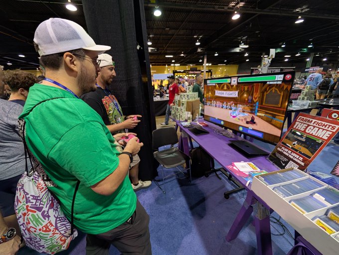 Two men playing a video game at a convention booth, with a screen displaying 'FARM FIGHTS THE GAME' and a promotional sign beside the monitor.