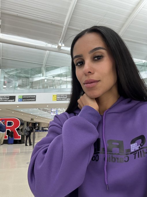 A woman with long dark hair wearing a purple hoodie takes a selfie in what appears to be an airport terminal, with a large red 'R' sign and airport signage visible in the background.