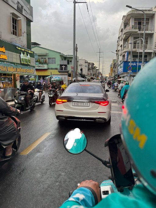 A first-person view from a motorcycle rider in teal Grab gear on a busy street, following a silver car with license plate '51G-111.11' through a Vietnamese city.