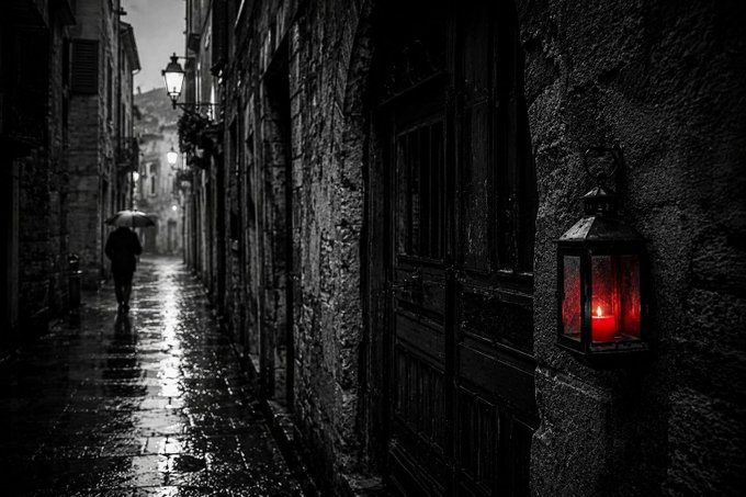A black and white atmospheric photo of a rainy, narrow cobblestone street with a single red lantern providing the only color on the right side.