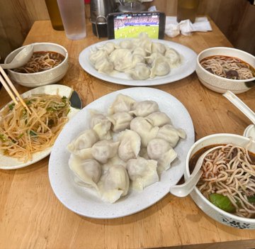 Two plates piled high with steamed dumplings are served alongside bowls of noodles and stir-fried vegetables at what appears to be a cozy restaurant, with "Sports" visible on a TV screen in the background, capturing the quintessential comfort food experience that suggests "SF doesn't miss."