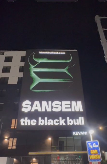 A nighttime photo of a large illuminated billboard on a building advertising 'blackbullsol.com' with '$ANSEM' and 'the black bull' in white text, featuring a green abstract design.